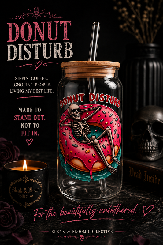 A clear glass jar tumbler with a bamboo lid and metal straw, featuring a colorful graphic of a skeleton reclining on a pink donut. The tumbler is set against a dark, moody background with a lit candle, a skull, and gothic text reading 'DONUT DISTURB'.