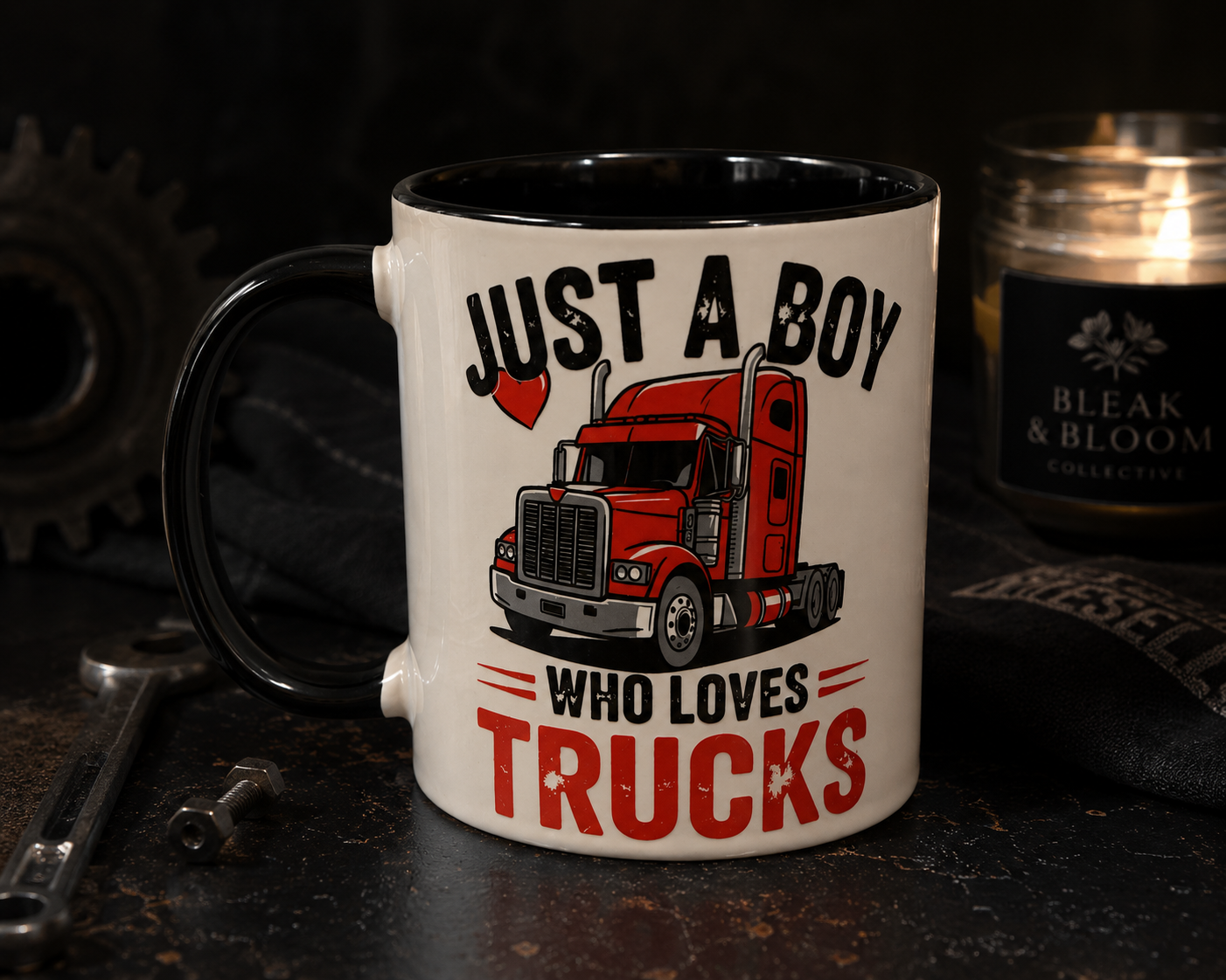 A white ceramic coffee mug with a black handle and interior, printed with the text 'JUST A BOY WHO LOVES TRUCKS' and an illustration of a red semi-truck. The mug is set on a dark rustic surface with a wrench, gear, and a lit candle in the background.