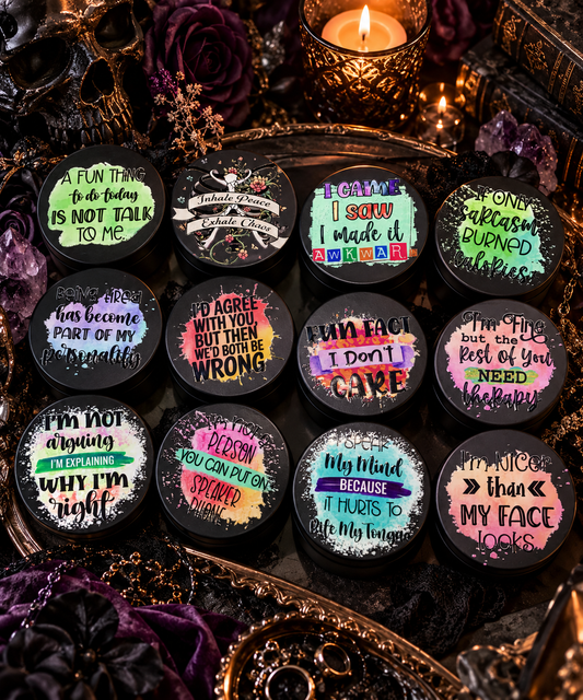 Twelve small black round containers with colorful text-based designs are arranged on a decorative tray with candles, crystals, and dark floral props. The containers feature various gothic and humorous quotes on pastel and neon backgrounds.