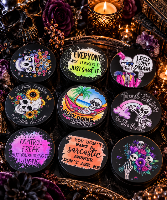 Nine round black cosmetic tins arranged in a flat lay with gothic props including candles, crystals, and a decorative tray. Each tin has a different colorful skull-themed graphic and humorous text such as 'Everyone was thinking it just said it' and 'Buy doing nothing.